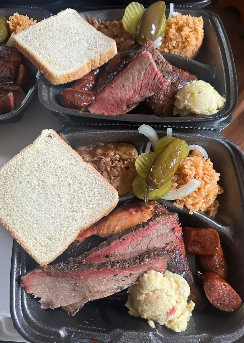 Two takeout barbecue plates with brisket, chicken, sausage, rice, beans, potato salad, bread, and pickles