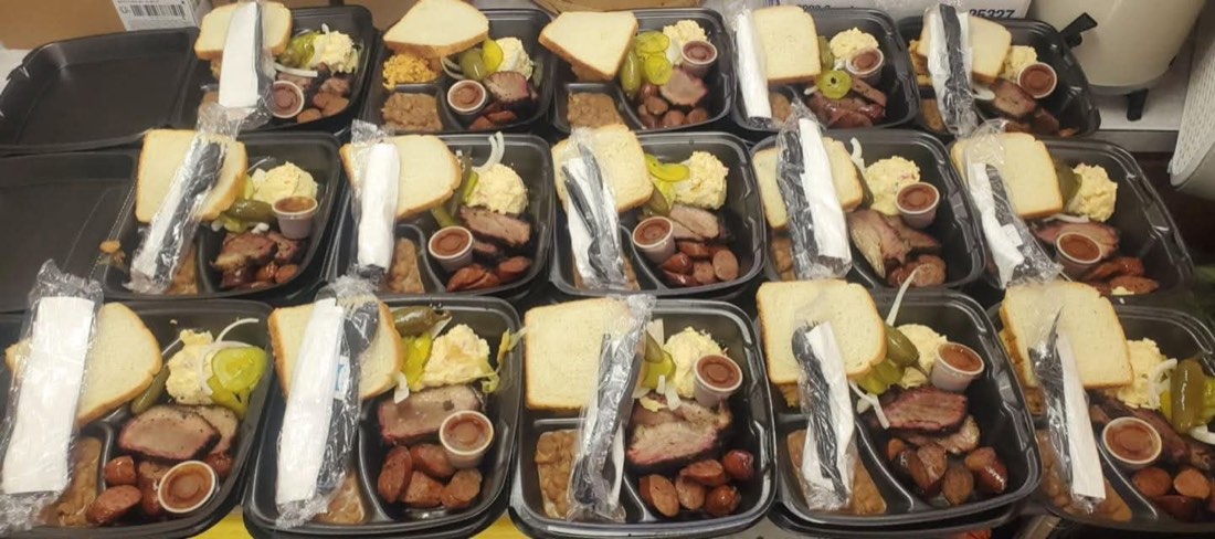 Rows of prepared barbecue meals with brisket, sausage, sides, bread, and sauce