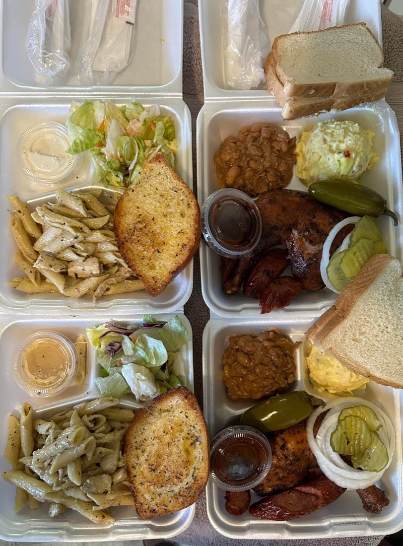 Four takeout plates with pasta, salad, smoked meats, beans, potato salad, bread, and pickles